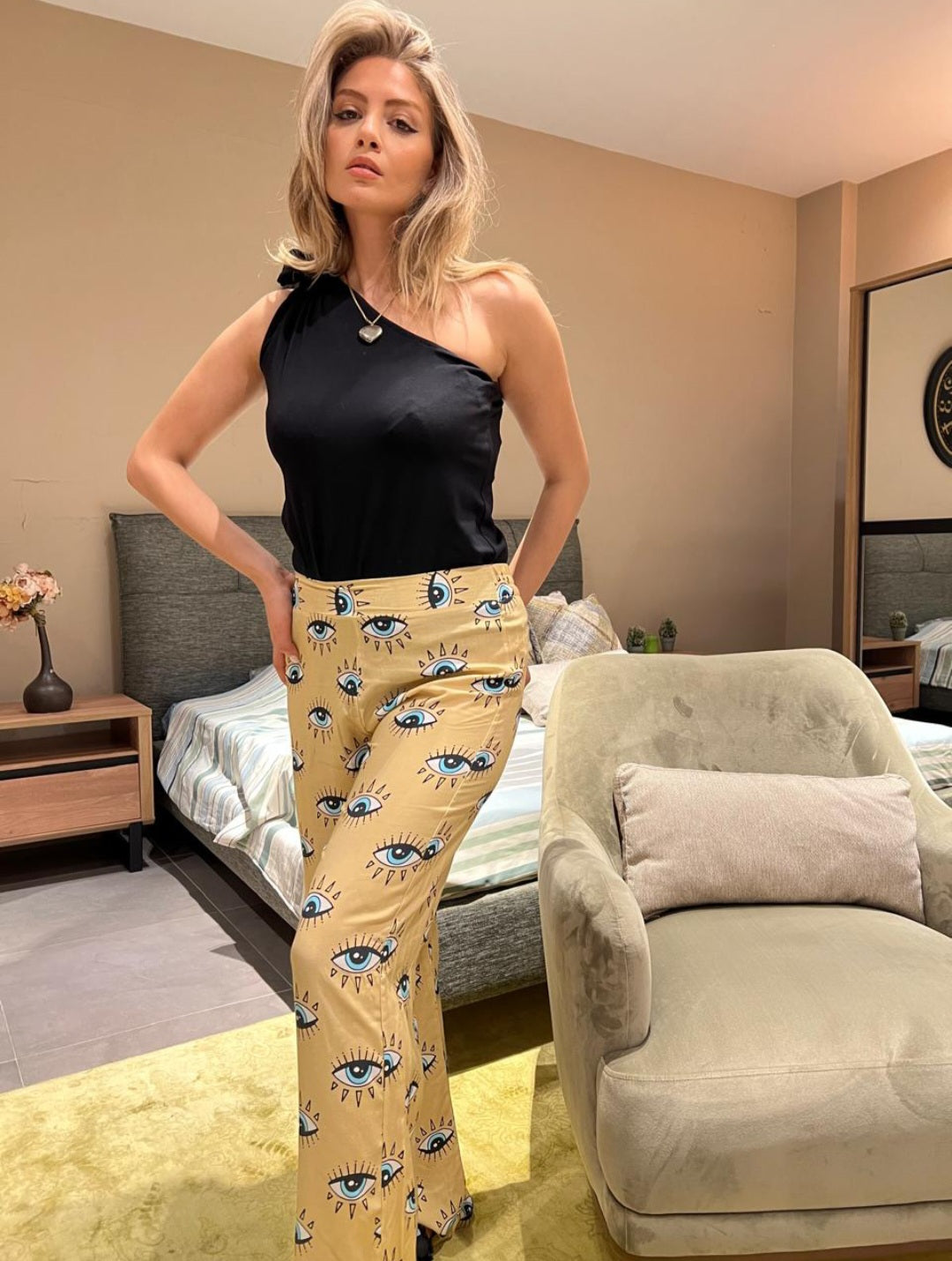 Woman wearing a trendy one-shoulder black top and beige pants with an eye pattern pyjama set by Popjammies, posing confidently in a modern bedroom.