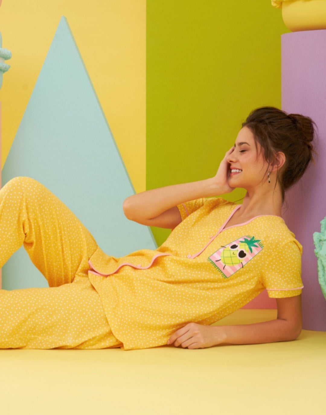 Model lounging in the Popjammies "Pineapple Delight" Pajama Set, featuring a vibrant yellow top with a playful pineapple graphic and matching yellow polka dot pants, posing against a colorful pastel backdrop.