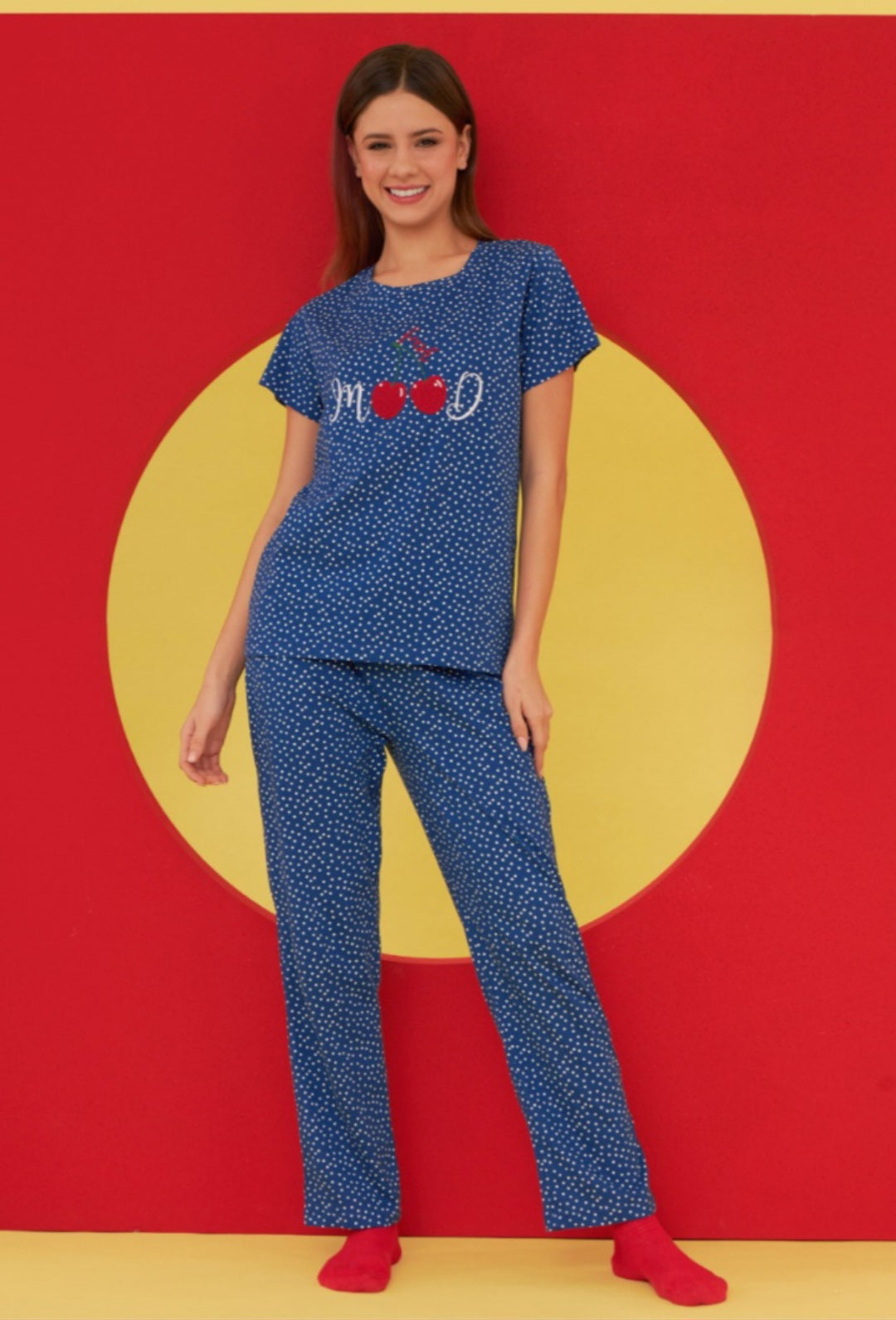 Model wearing the Popjammies "Cherry Mood" Pajama Set, which includes a blue top with a cherry motif and the word "MOOD" in script, paired with matching blue dotted pants, standing against a colorful backdrop.