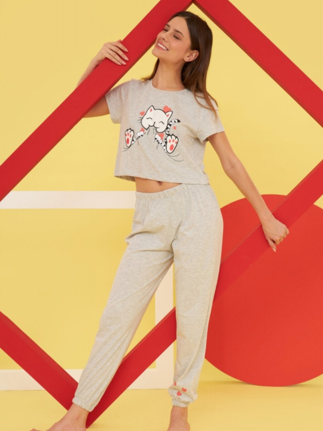 Model posing in the Popjammies Cozy Cat Pajama Set, smiling with closed eyes. The set includes a light grey cropped top with a cute cat graphic and matching jogger pants with paw print details. The vibrant yellow and red background adds a playful touch, making the cozy loungewear stand out.