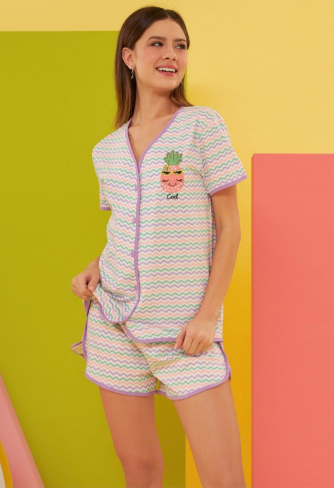 A model wearing the Pineapple Zigzag Pajama Set, featuring a short-sleeve button-up top and matching shorts with a fun zigzag pattern in pastel shades and an adorable pineapple graphic with sunglasses.