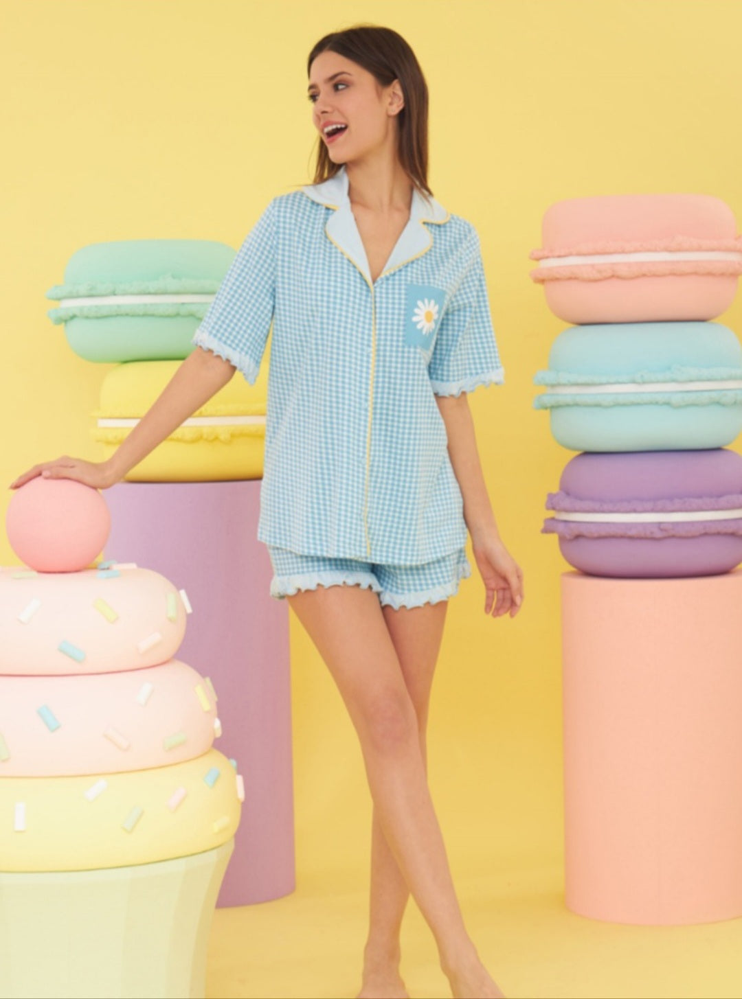 A model wearing the  Blue Gingham Pajama Set with a daisy pocket detail and yellow piping. The set includes a short-sleeved button-up top and matching ruffled shorts, perfect for stylish and comfortable sleepwear. 