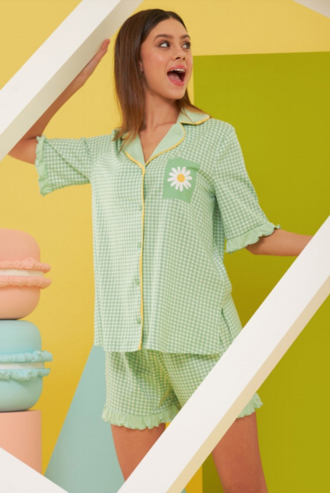 A model wearing a green gingham pajama set with daisy pocket detail and yellow piping, featuring a button-up shirt and matching ruffled shorts.