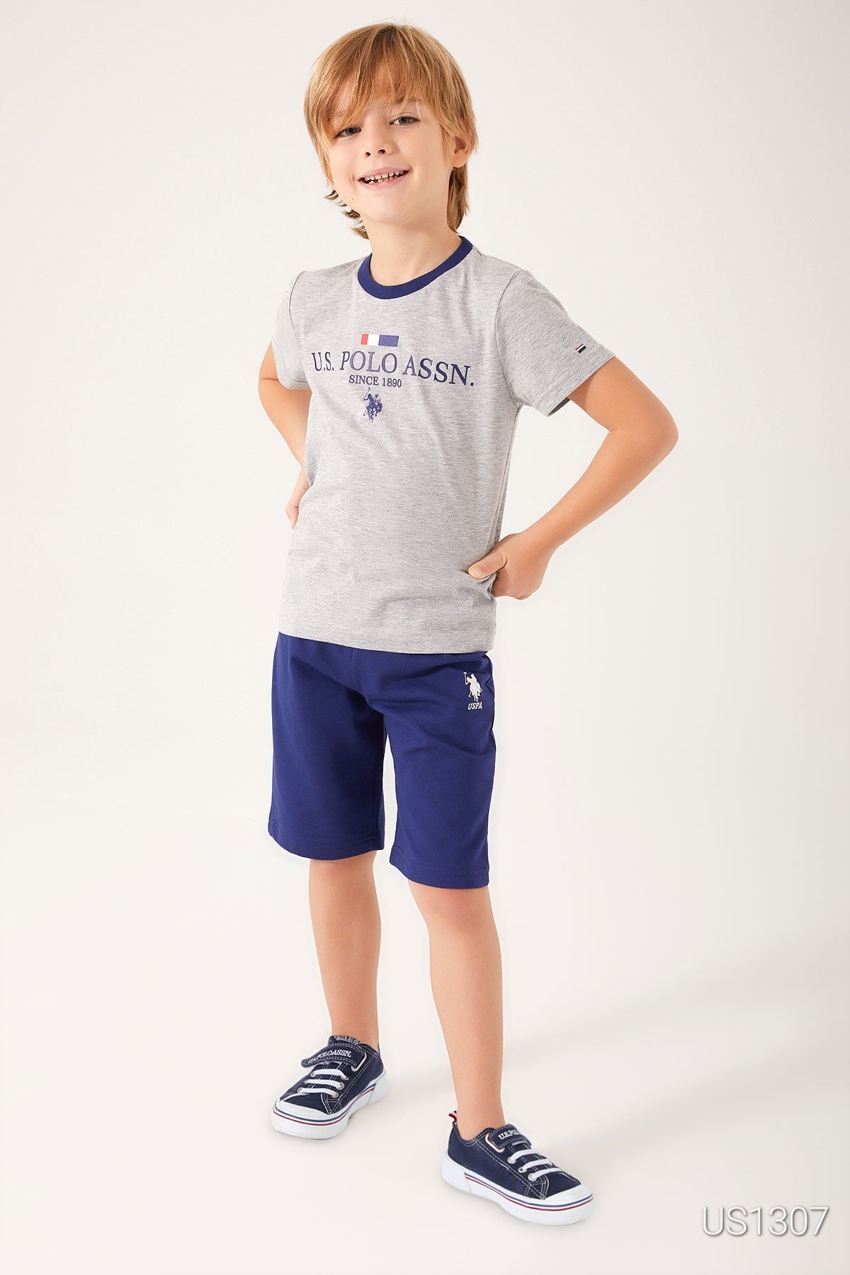 Front view of boy wearing grey top with "US Polo Assn" and navy blue shorts, smiling.