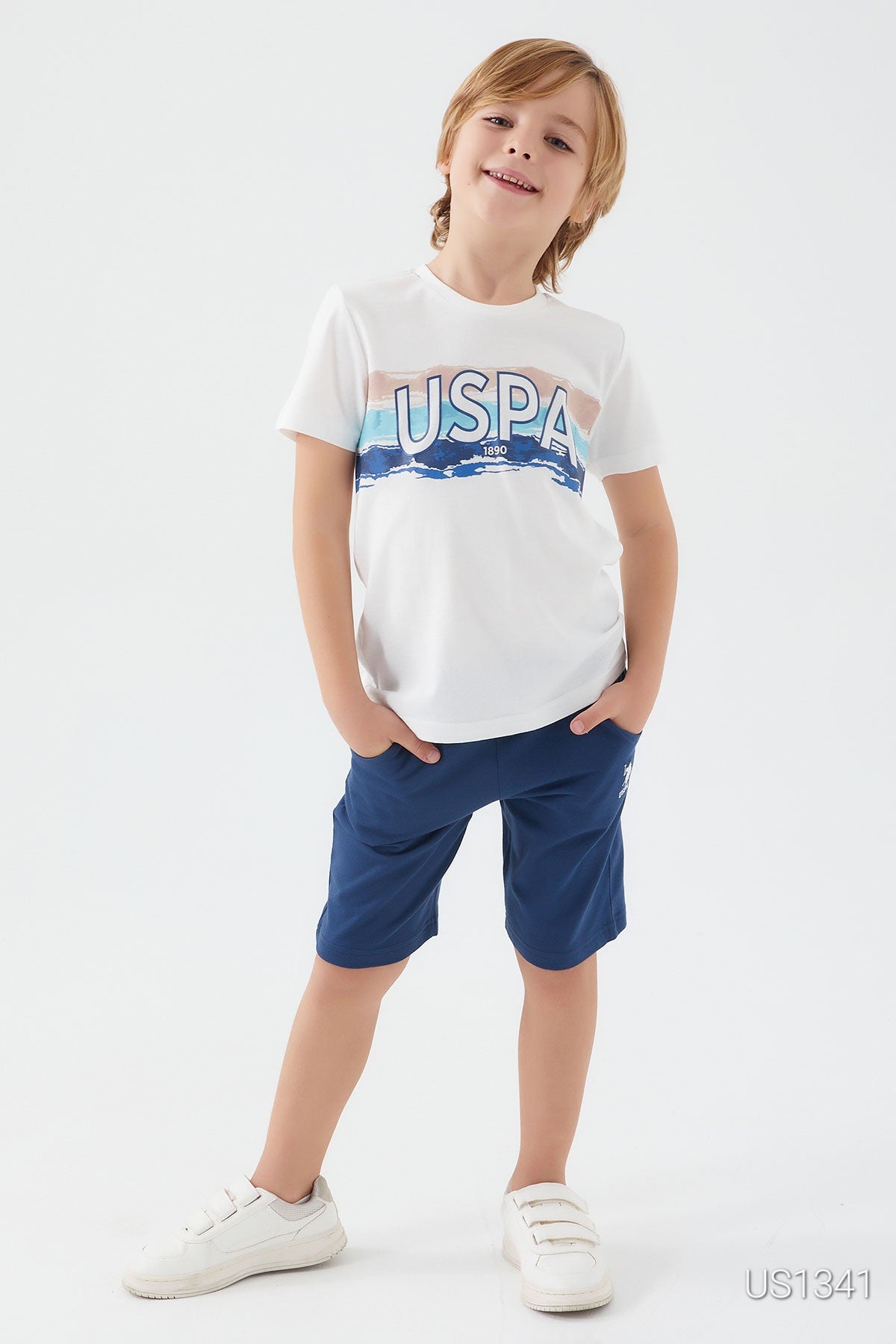  Boy wearing a white top with "USPA" graphic and navy shorts, smiling with hands in his pockets.