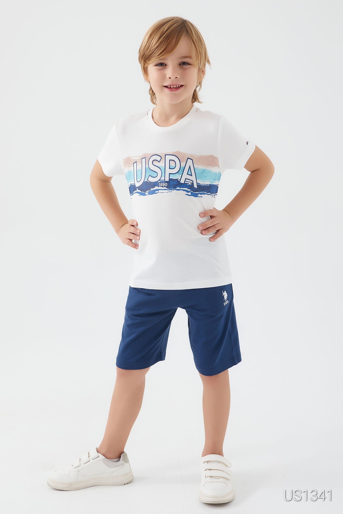  Boy wearing a white top with "USPA" graphic and navy shorts, smiling with hands on hips.