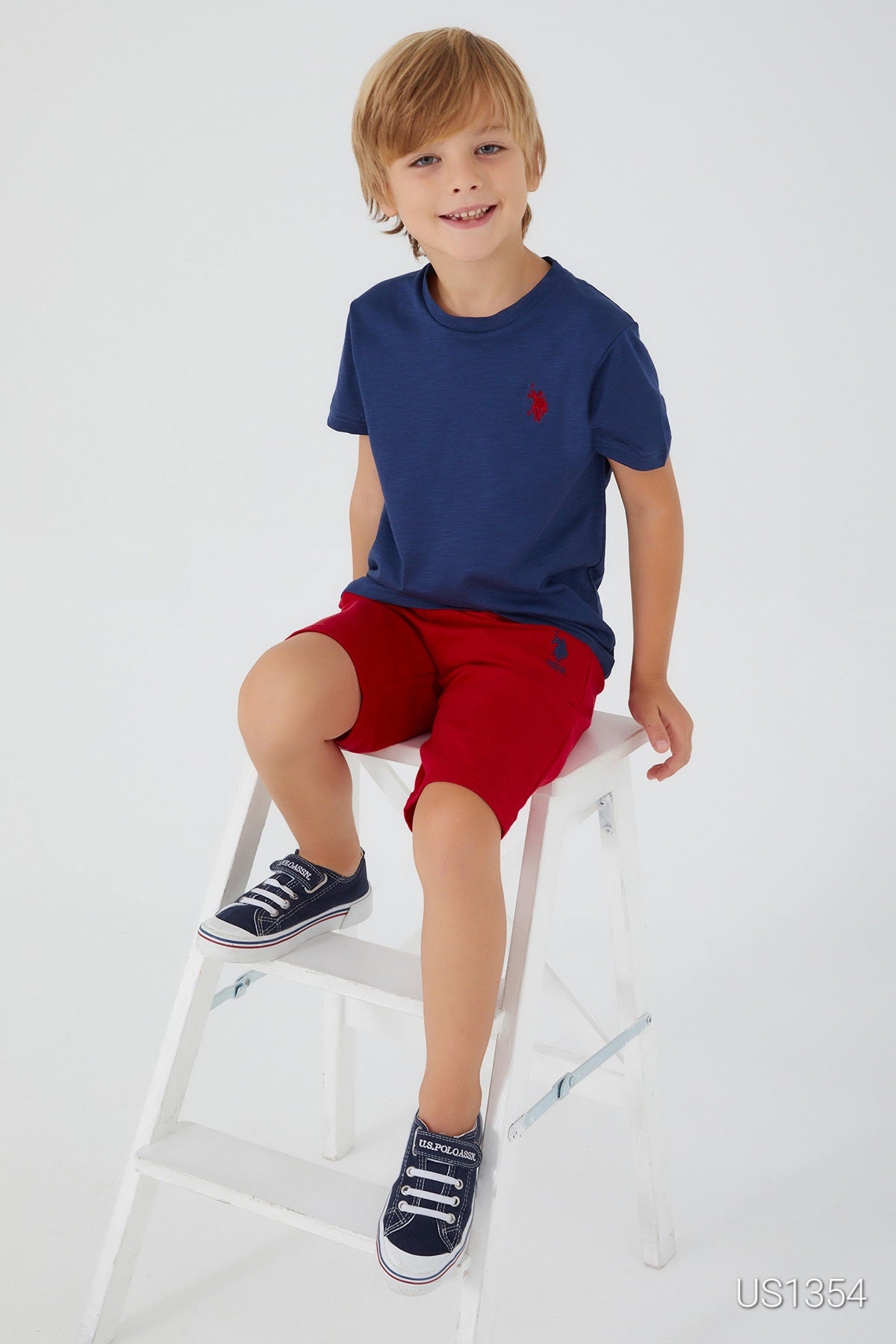 A boy sitting on a white chair wearing a navy top with bold red "USPA" lettering and matching red shorts.