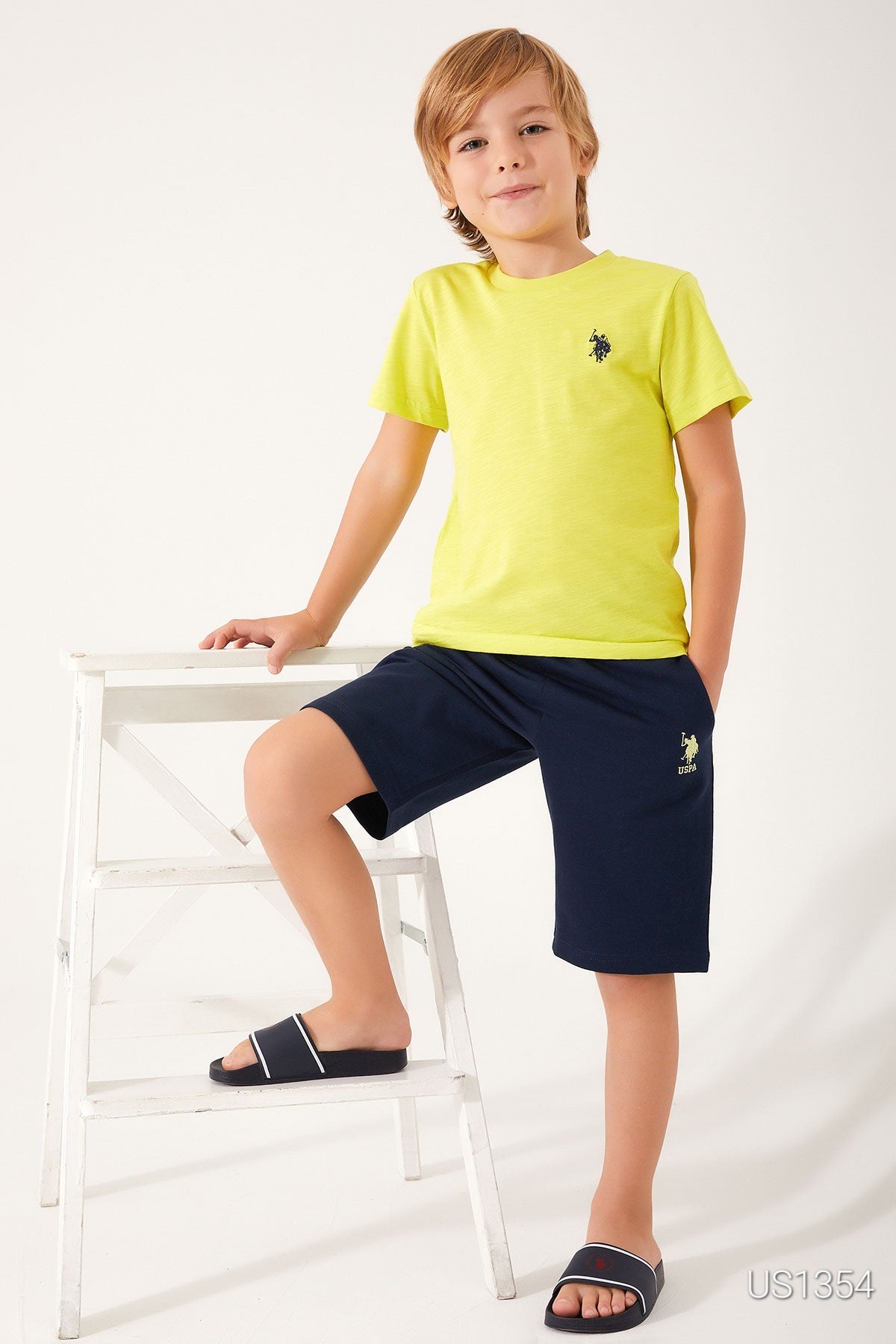 A boy posing beside a white chair wearing Popjammies Yellow USPA Top and Navy Shorts