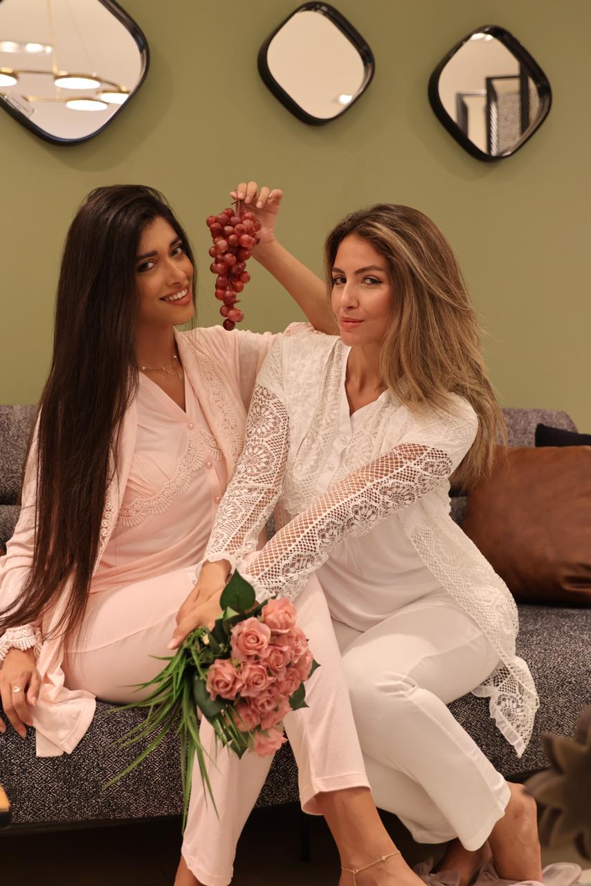 Two women in Popjammies Lace Trim Maternity Pajama Set, one in pink and one in white, sitting on a couch with grapes and flowers.