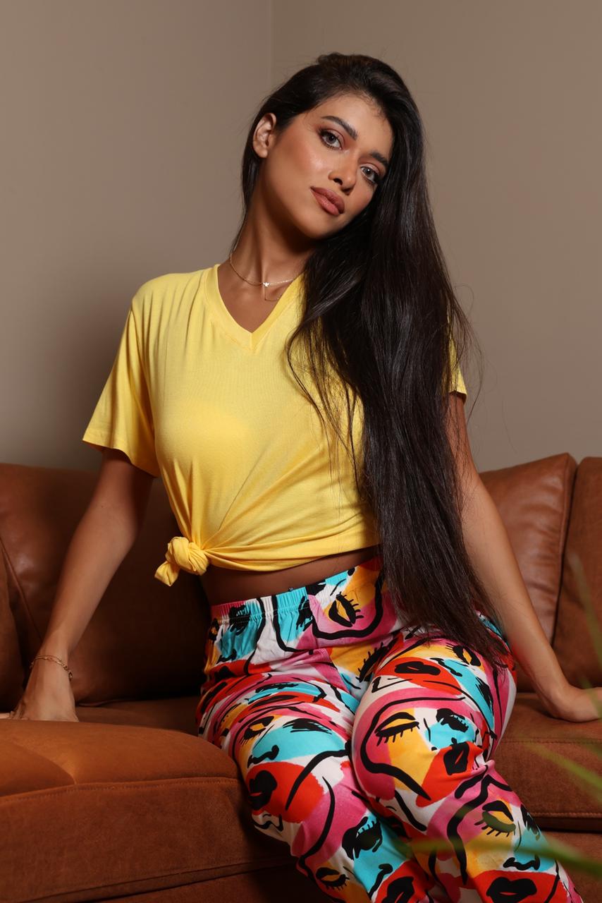 Model wearing Popjammies Abstract Art Pyjama Set with a vibrant multicolored abstract design and a yellow V-neck tee, sitting on a brown sofa.