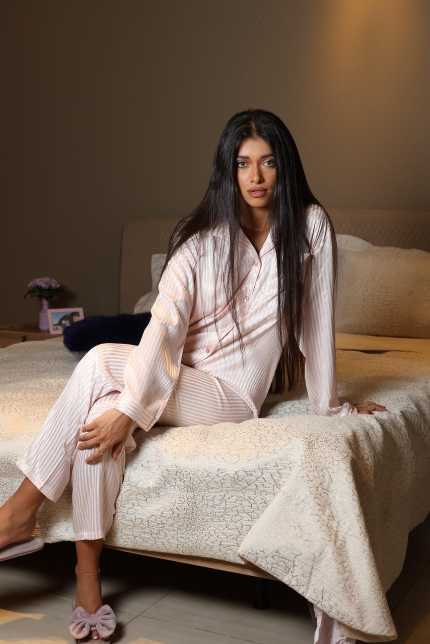 Model sitting on a bed wearing a pink striped silky pyjama set by Popjammies, with slippers and gazing ahead.