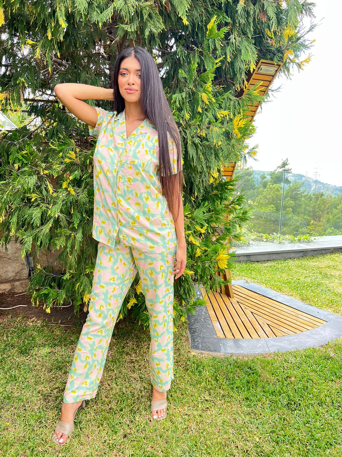 Woman standing outdoors in front of a green tree, wearing a light green and pink patterned pyjama set with short sleeves and long pants.