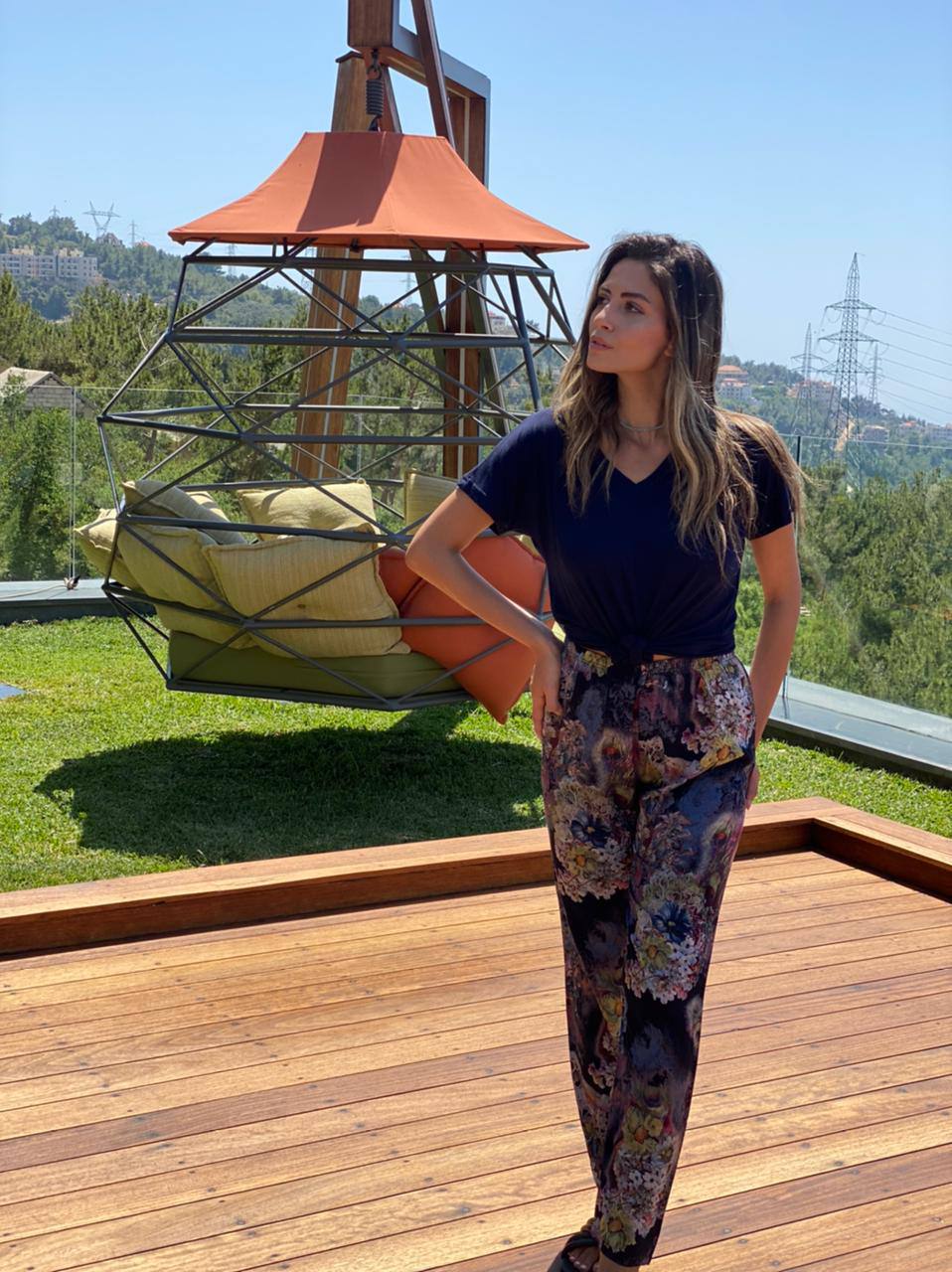 Woman in navy blue top and floral print pants by Popjammies, standing on a wooden deck with a hanging chair in the background.