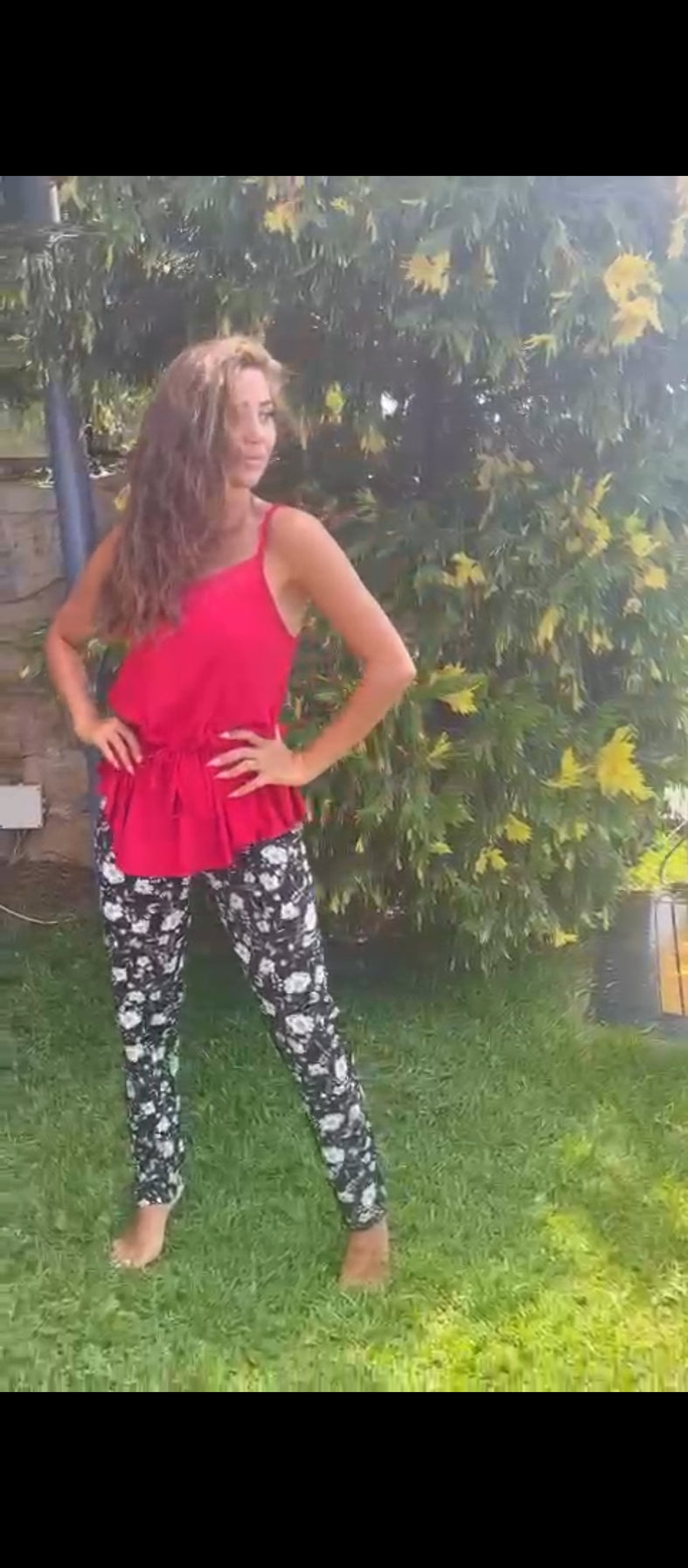 Model wearing a red top and black floral print pyjama pants by Popjammies, standing confidently outdoors with a background of green foliage.