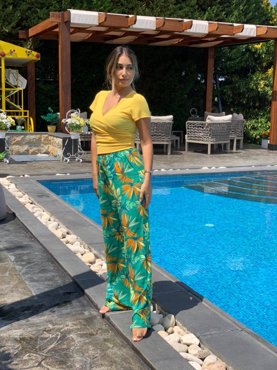 Woman standing by the poolside wearing a yellow top and green floral Popjammies pyjama pants.