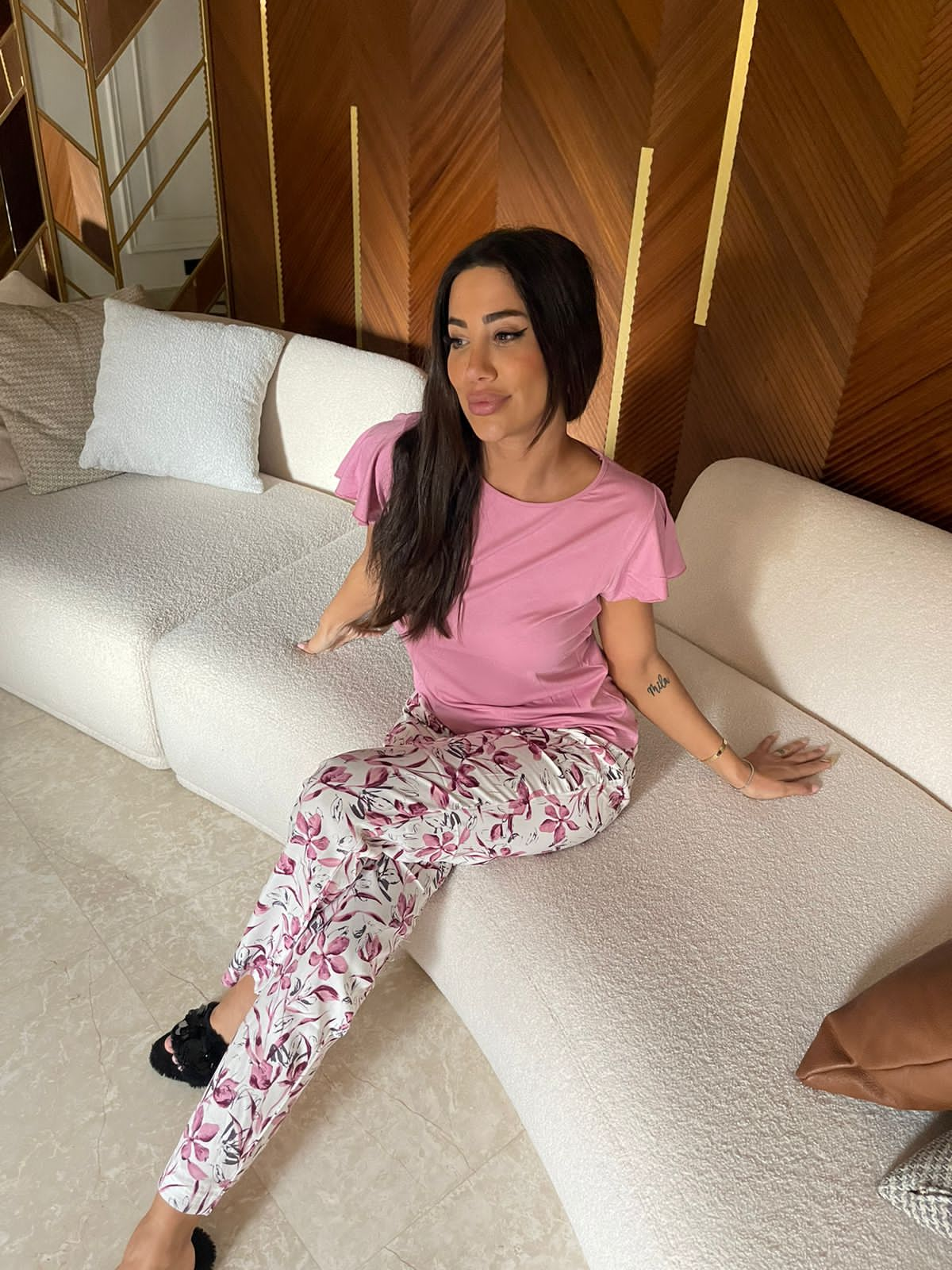 Model wearing a Popjammies lilac flutter sleeve pyjama set, featuring a soft lilac top and white pants with a pink and navy floral print, seated on a beige couch with a wooden background.