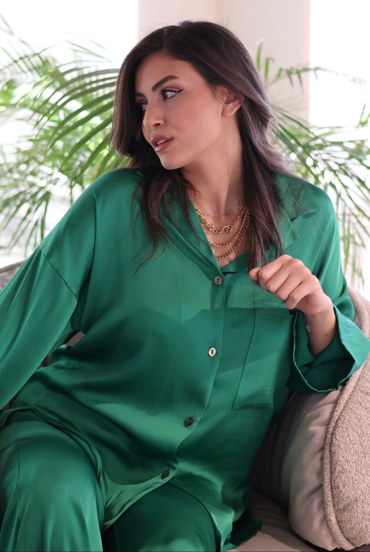 Woman in a green silk pyjama set by Popjammies, sitting on a couch with plants in the background.