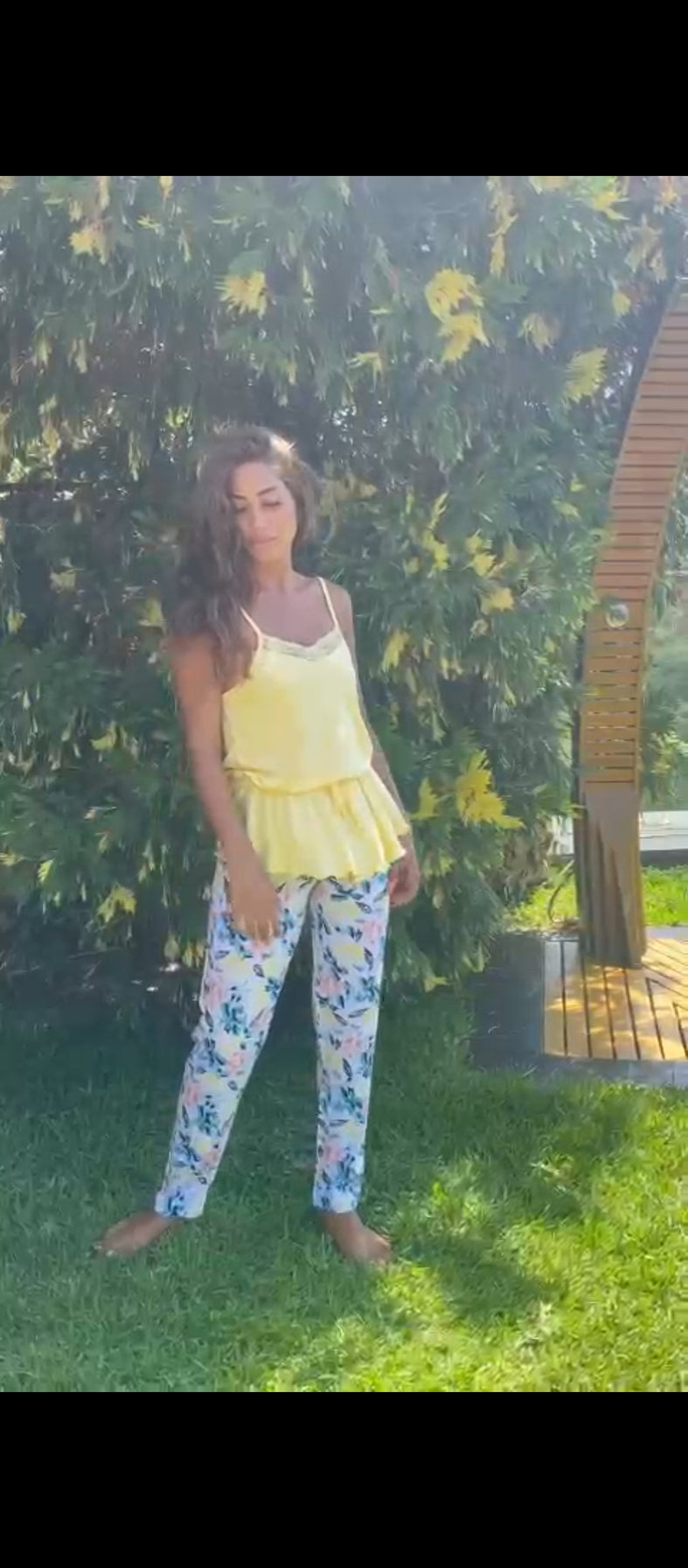 Model wearing a yellow top and white floral print pyjama pants by Popjammies, standing in a sunny garden with green foliage in the background.