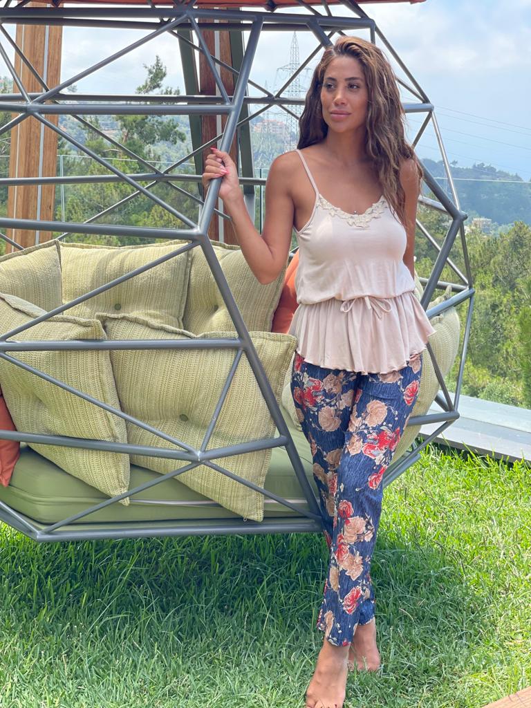 Model wearing a floral print pyjama set by Popjammies, featuring a beige top and floral print navy pants, standing outdoors next to a geometric chair with a scenic background.