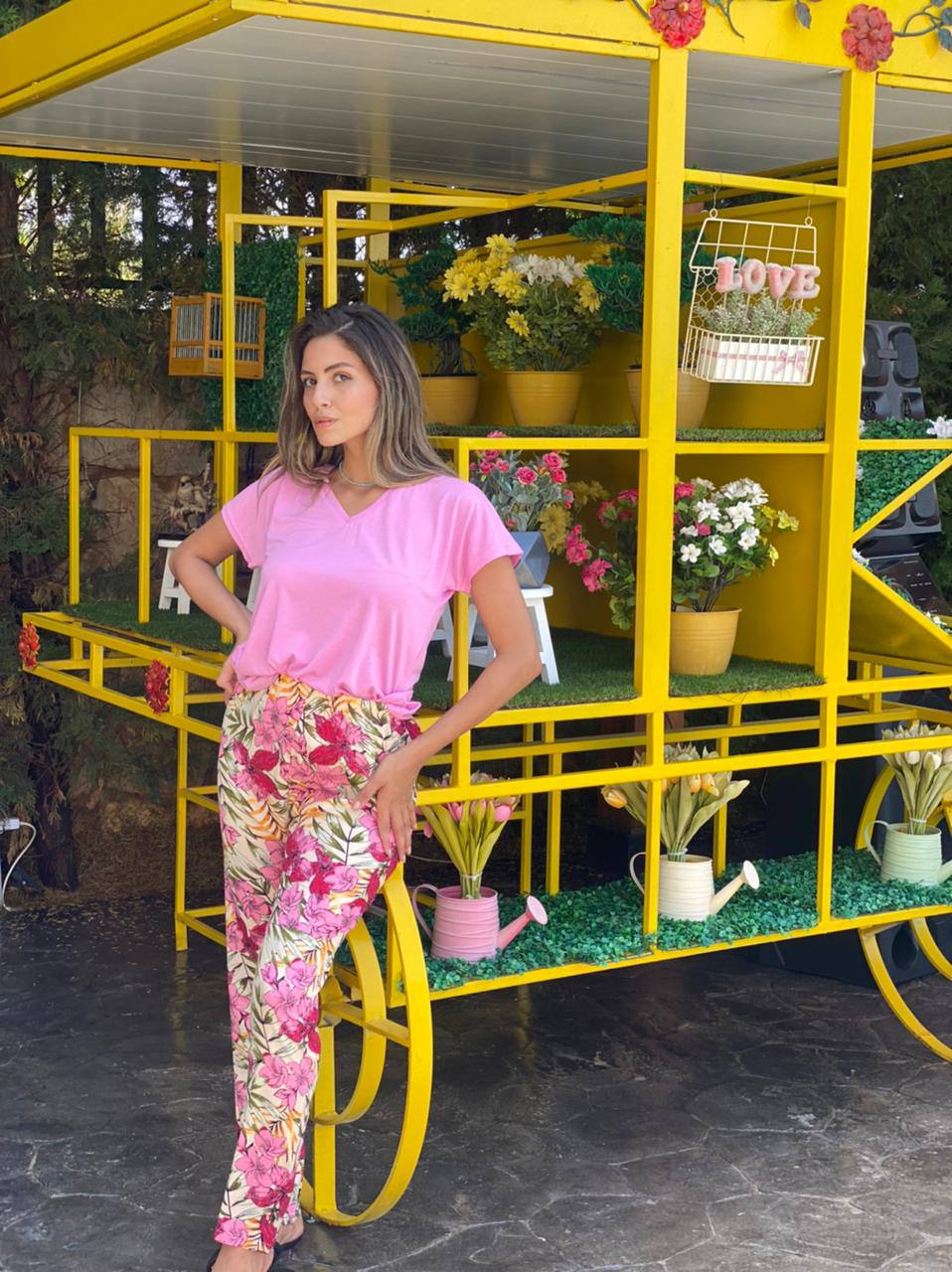 Woman in pink top and floral print pants by Popjammies, standing in front of a yellow cart with potted flowers.