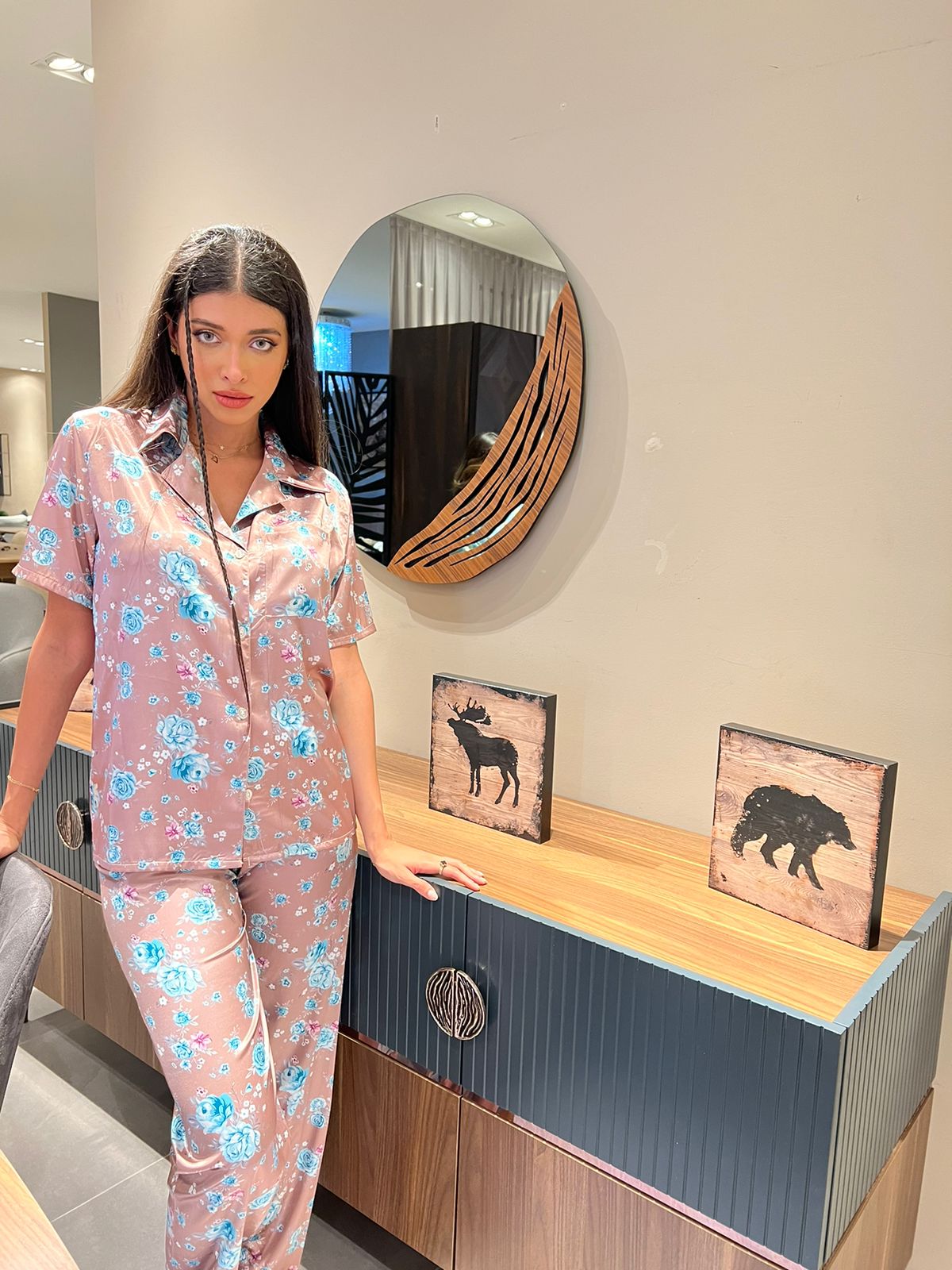 Woman in pink and blue floral satin pajamas, standing next to a modern wooden dresser.