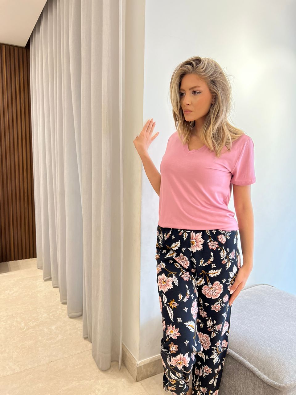 Woman standing beside a curtain wearing a pink V-neck T-shirt and black pajama pants with a floral pattern.