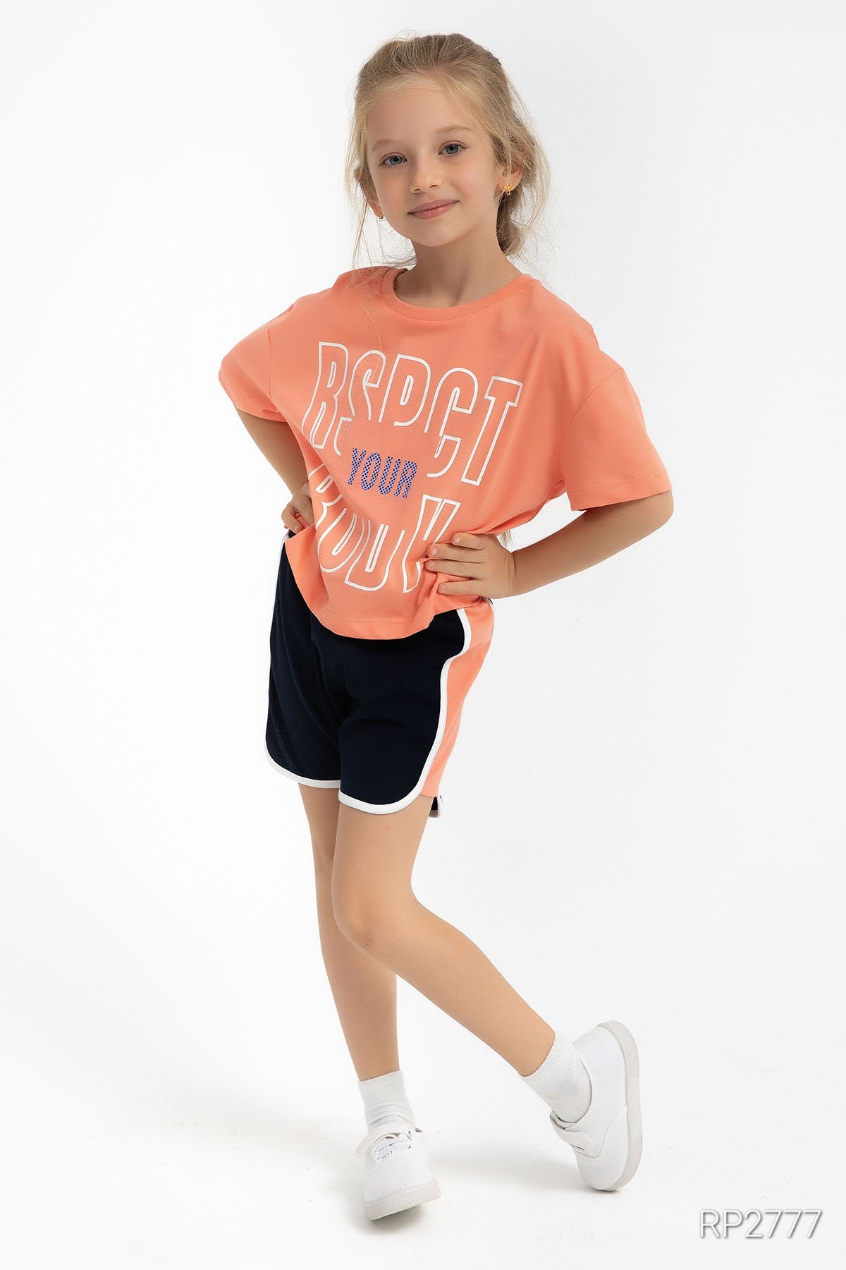 Girl wearing an orange top with black shorts in front of a plain background.