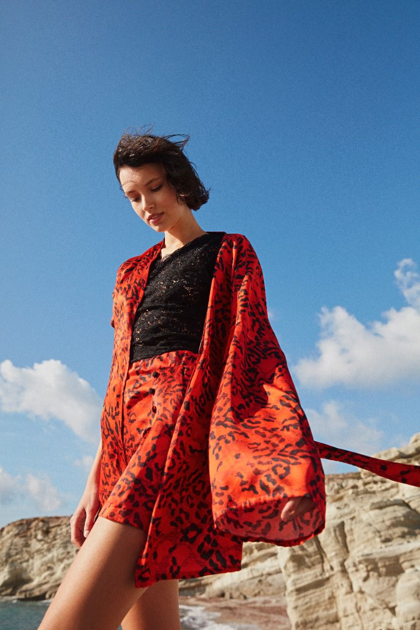 Model wearing a Popjammies red and black leopard print pyjama set, including a robe, matching shorts, and a black lace top, standing outdoors with a clear blue sky in the background.