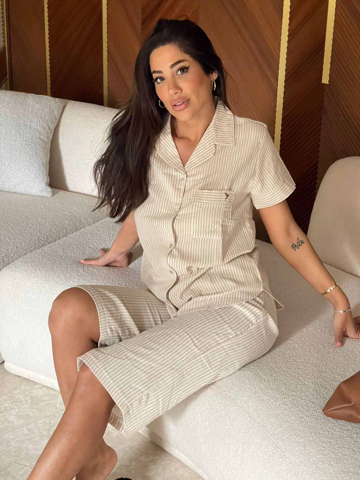 Woman sitting on a sofa wearing a beige short-sleeve button-down top with vertical stripes and matching capri pantacour