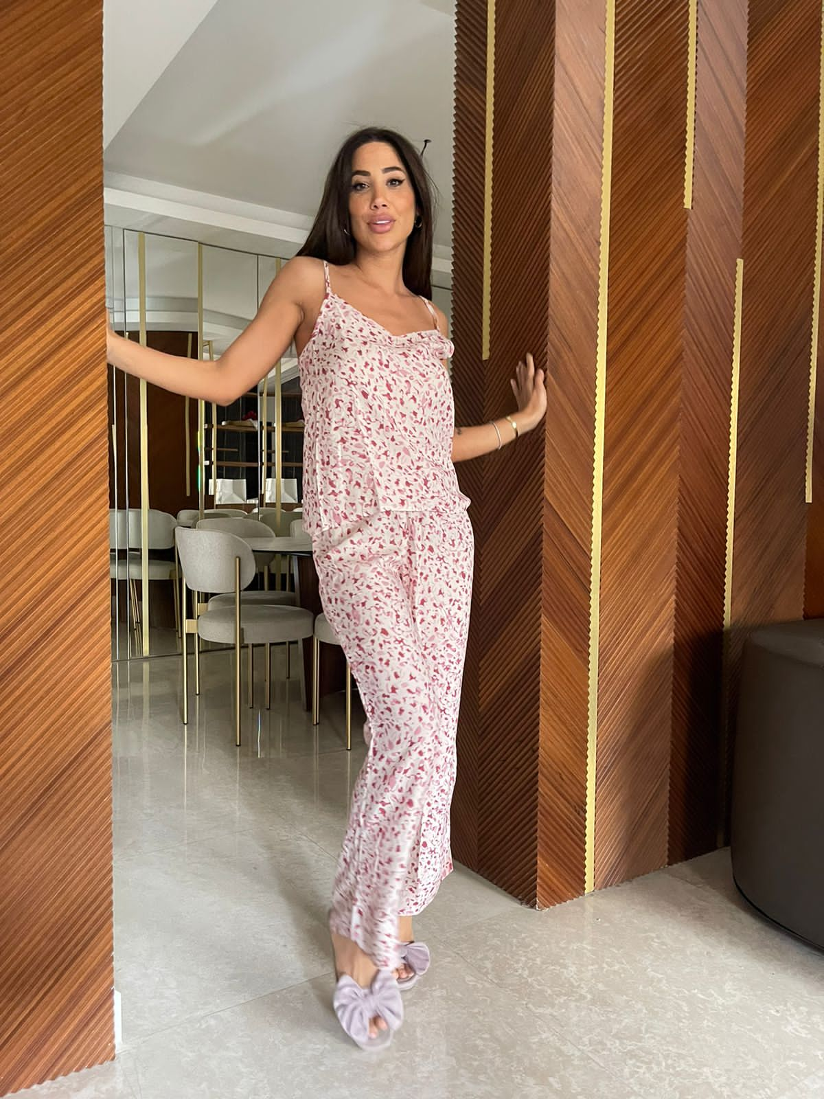 Woman in a pink floral sleeveless pajama set standing indoors, leaning slightly with one arm resting against a wood-paneled wall, with a dining area in the background.