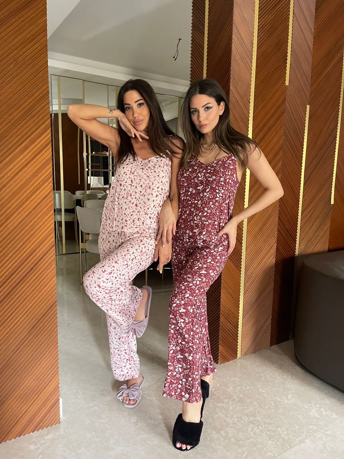 Two women are posing in a modern room with wooden panel walls. The woman on the left is wearing a pink and white floral spaghetti strap pajama set, while the woman on the right is wearing a red floral spaghetti strap pajama set.