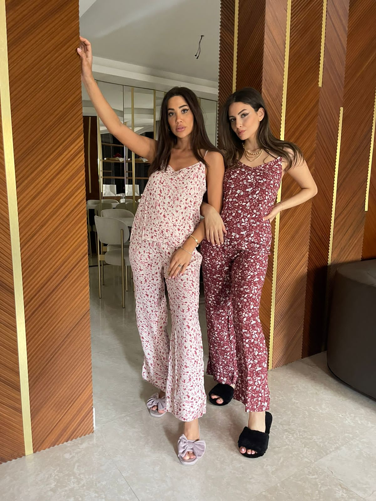 Two women are standing in a modern room with wooden panel walls. The woman on the left is wearing a pink and white floral spaghetti strap pajama set, while the woman on the right is wearing a red floral spaghetti strap pajama set.