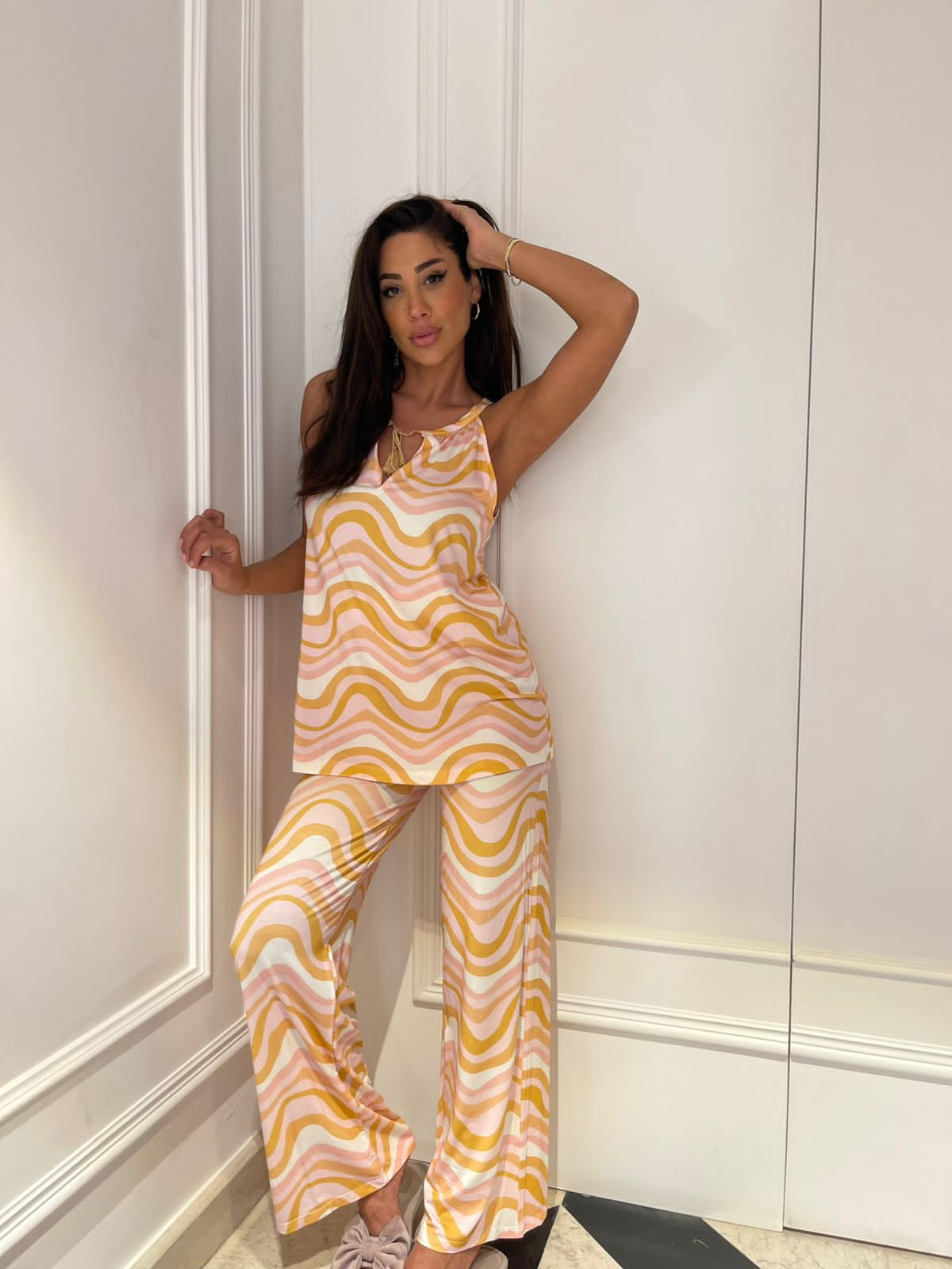 A woman posing against a white wall, wearing a pastel pink and yellow wave pattern pyjama set from Popjammies. She has long dark hair, one hand resting on her head, and is looking directly at the camera.