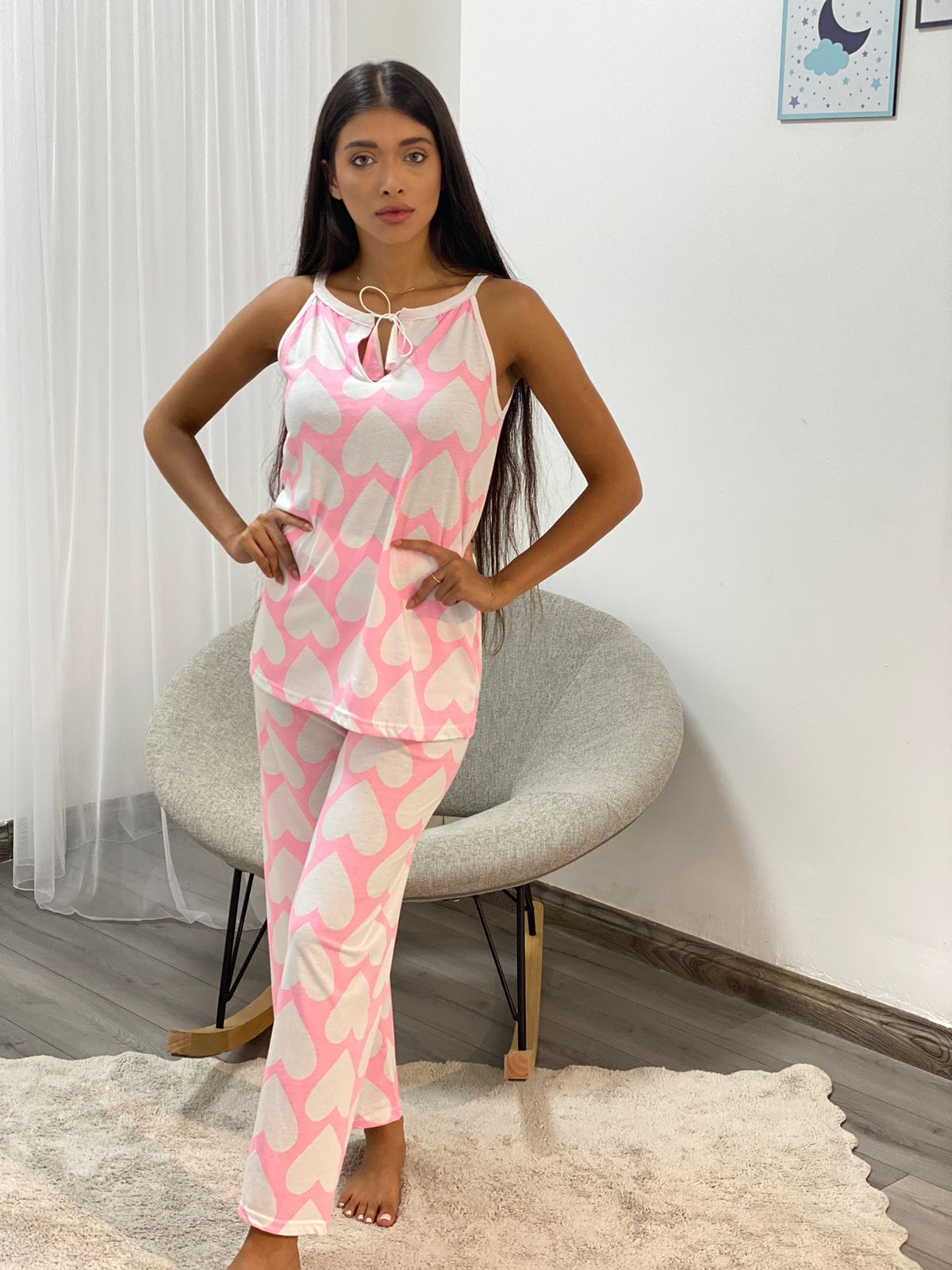 Woman wearing the Pink Heart Pajama Set by Popjammies, featuring a sleeveless top with bow tie at the neckline and matching pants, both with a large pink heart print on a white background.