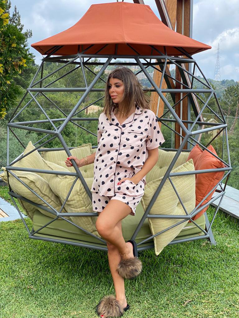 Woman wearing a pink polka dot pajama set from Popjammies, standing by a modern outdoor hanging chair with colorful cushions.