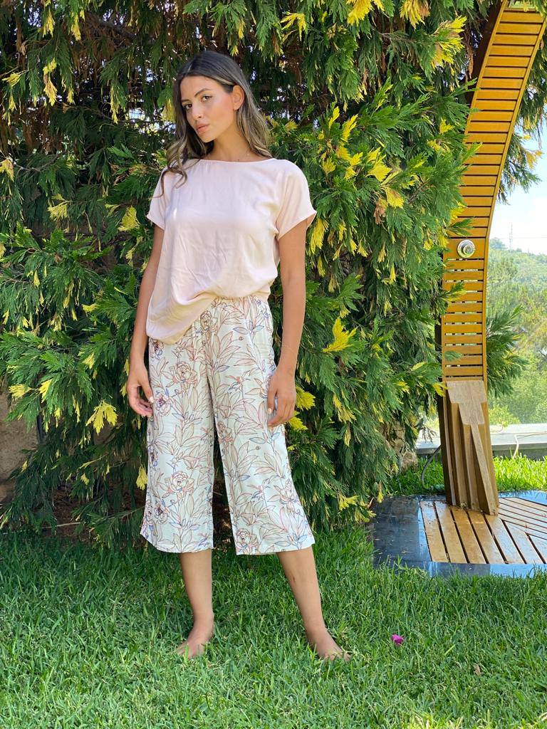 Woman wearing a light pink short-sleeve top paired with cream capri pantacour with a floral print from Popjammies, standing in a garden. 