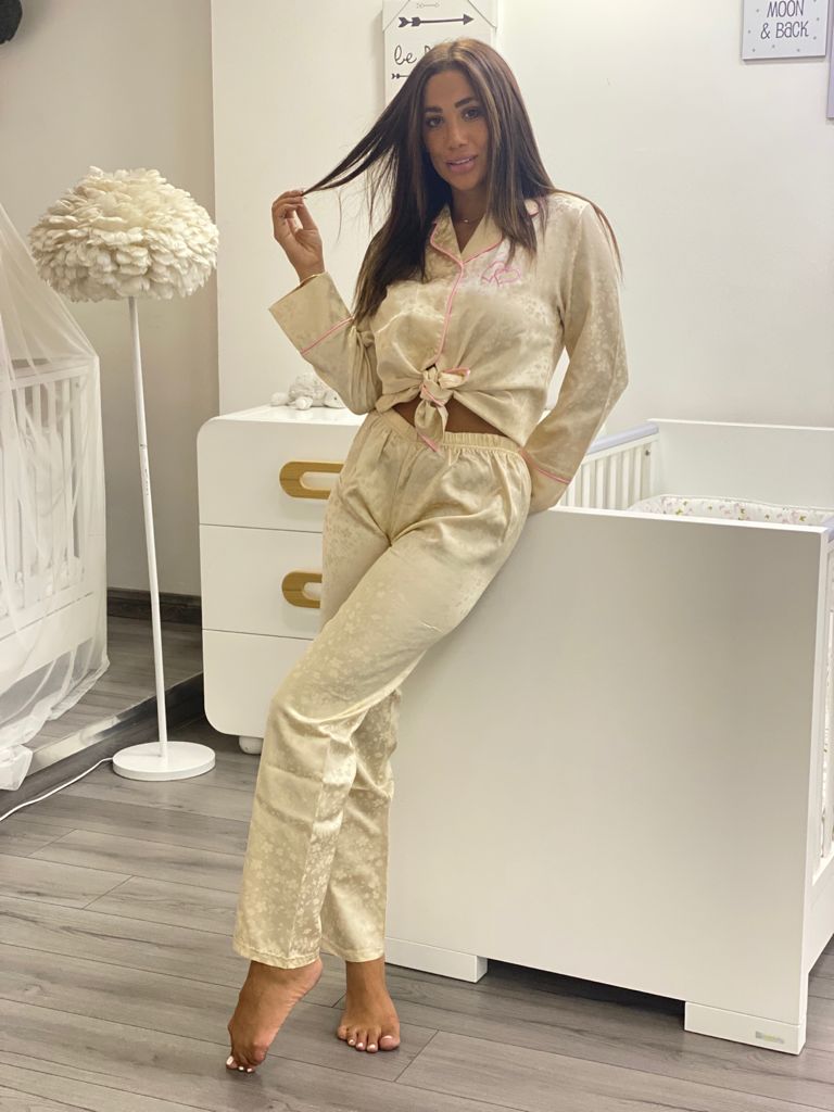 A woman standing in a nursery room, wearing a luxurious beige satin pajama set with pink piping, with the top tied at the waist, smiling and posing casually.