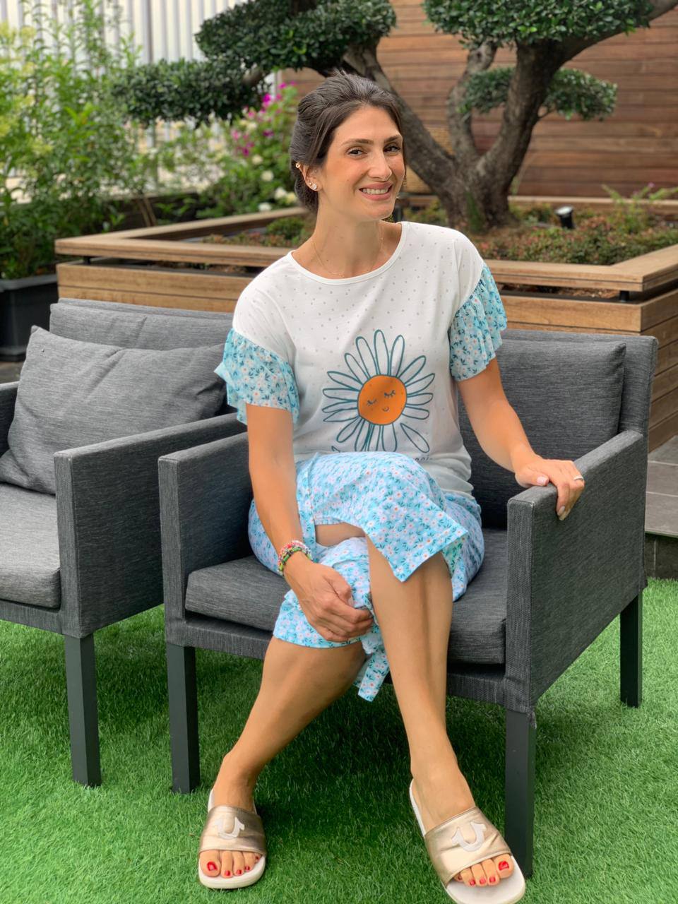 Woman wearing a white short-sleeve top with a daisy graphic paired with blue floral print capri pants from Popjammies, sitting in a garden.