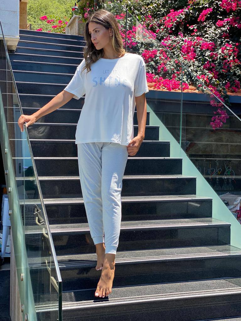 Woman wearing a white pajama set with a "Let It Shine" printed top and matching white striped pants, standing barefoot on outdoor steps with blooming flowers in the background.