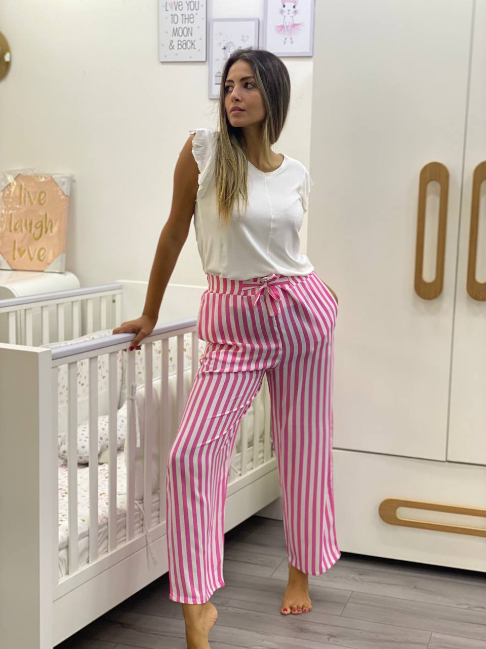 Woman wearing a white sleeveless top with ruffle sleeves and pink and white striped pajama pants, standing beside a white crib in a nursery room.