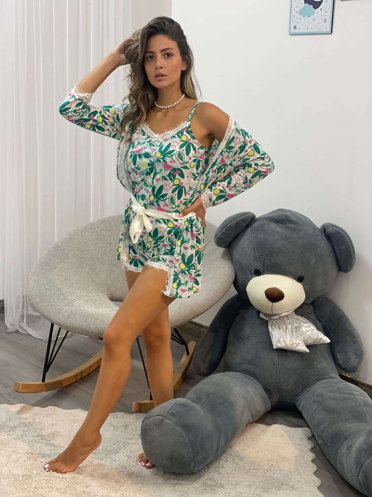 Model wearing a vibrant floral print pyjama set with lace trim and coordinating robe, standing confidently next to a large grey teddy bear in a modern, cozy room.