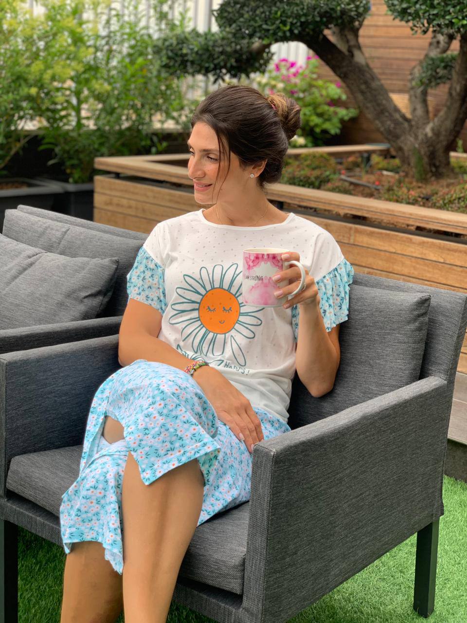 Smiling woman in a white daisy graphic top and blue floral print capri pants from Popjammies, enjoying a drink in a garden.