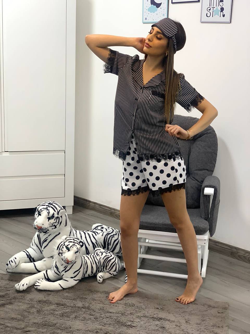 Woman posing in a black and white striped pajama set with polka dot shorts and matching sleep mask, next to stuffed tiger toys in a nursery room.